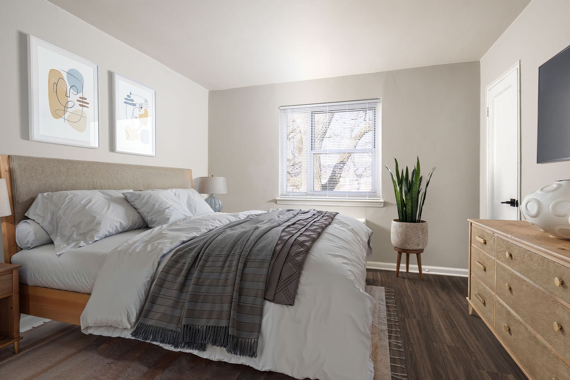 Bedroom in a neutral-toned apartment with a bed, dresser, plant, and a window.