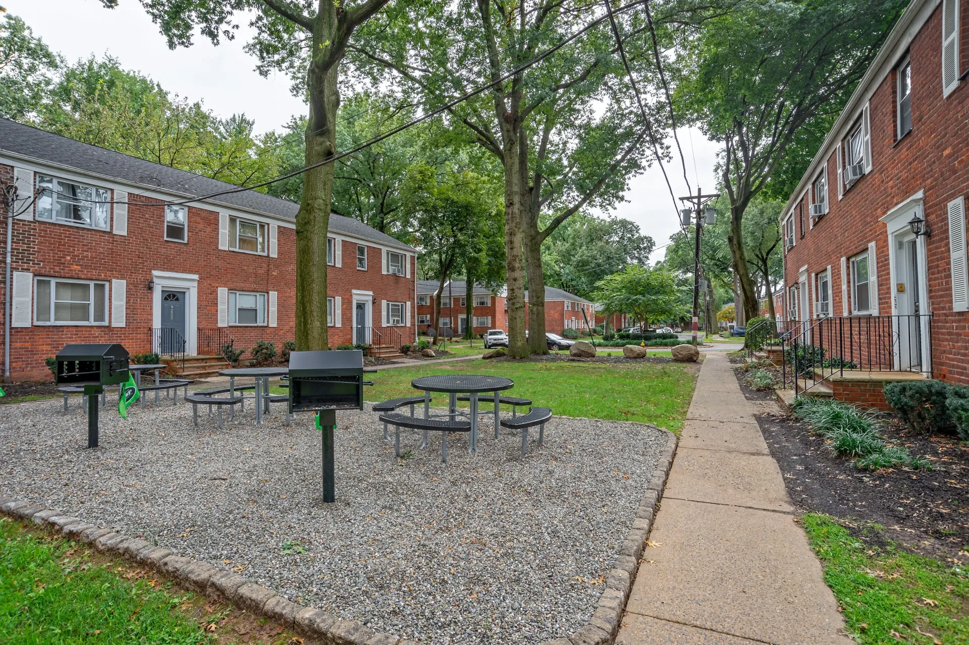 Outdoor community courtyard with brick apartment buildings, trees, gravel seating area, grills, and a central table.
