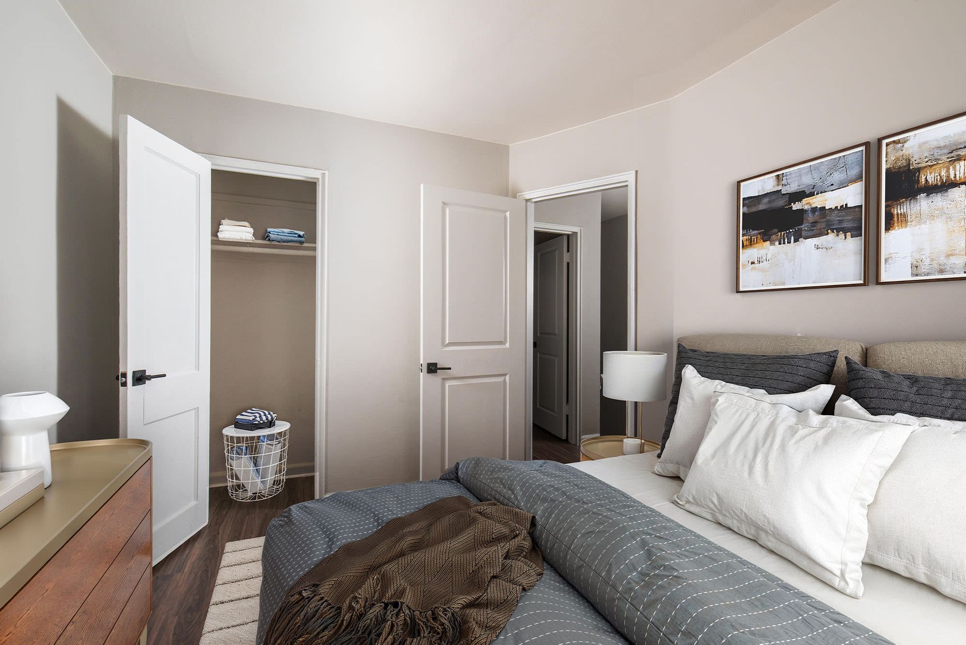 Bedroom in a modern apartment with a made bed, dresser, lamp, and open closet.