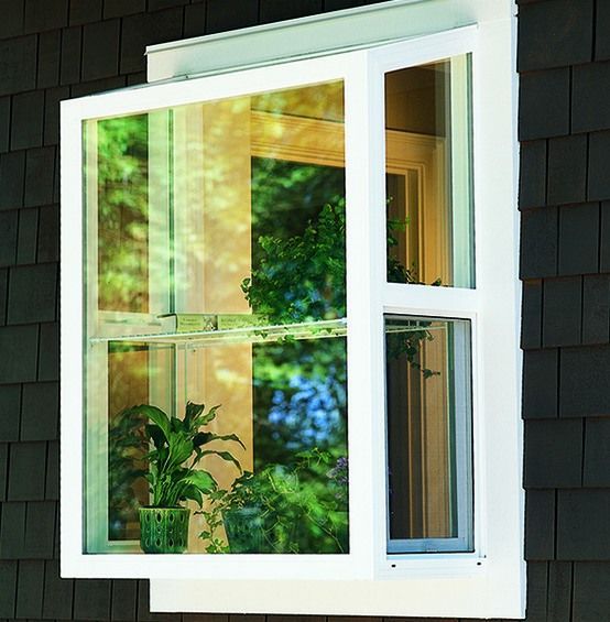 White-framed casement window, open, on dark siding. Reflects greenery and interior window.