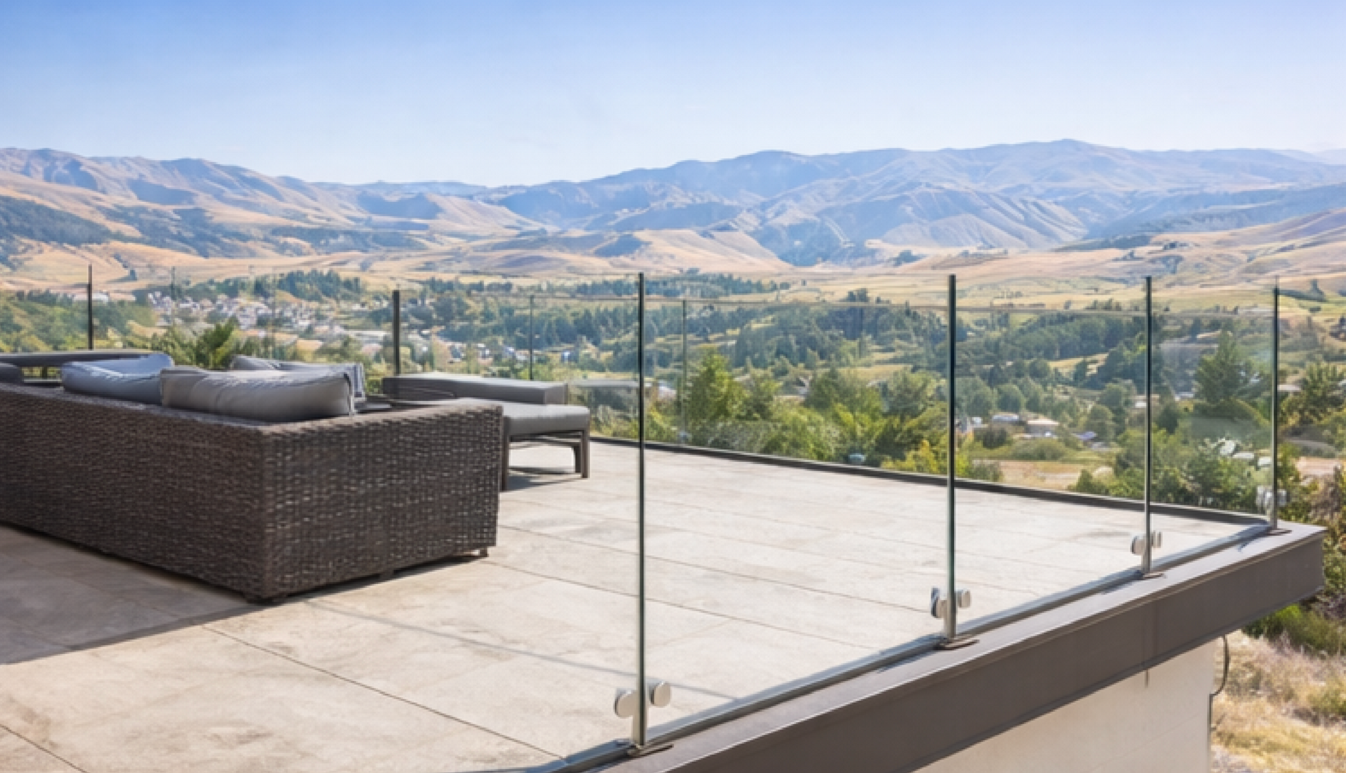 Rooftop patio with glass railing overlooking a valley and mountains. Brown seating. Sunny, clear sky.