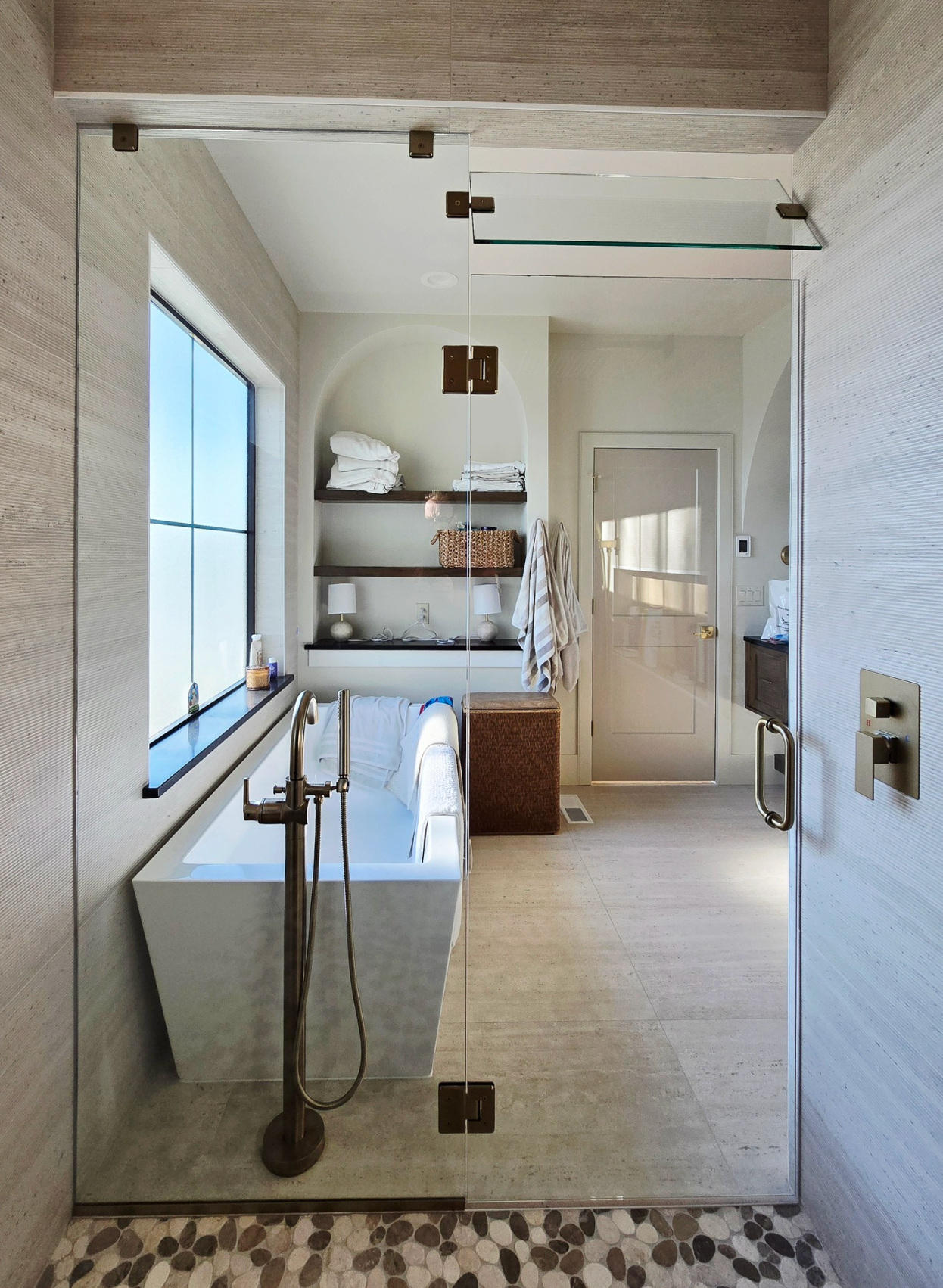 Modern bathroom: glass shower door, soaking tub, wooden shelves with towels, neutral colors.
