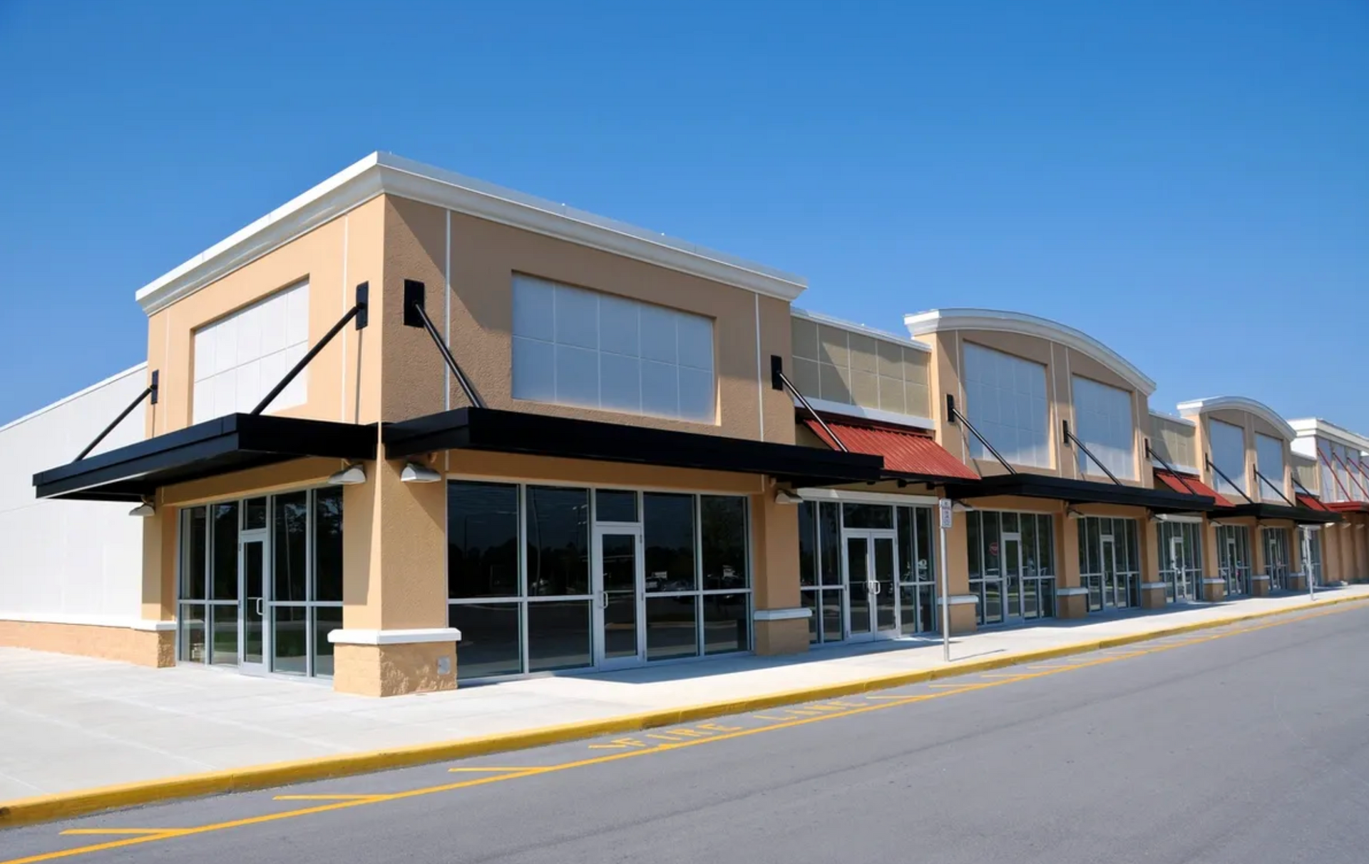 Empty beige commercial storefronts with black awnings under a bright blue sky.