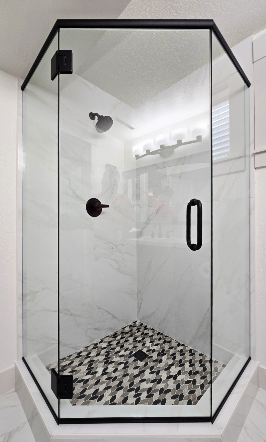 Glass shower enclosure with black trim and hardware, white marble-look walls, mosaic tile floor.