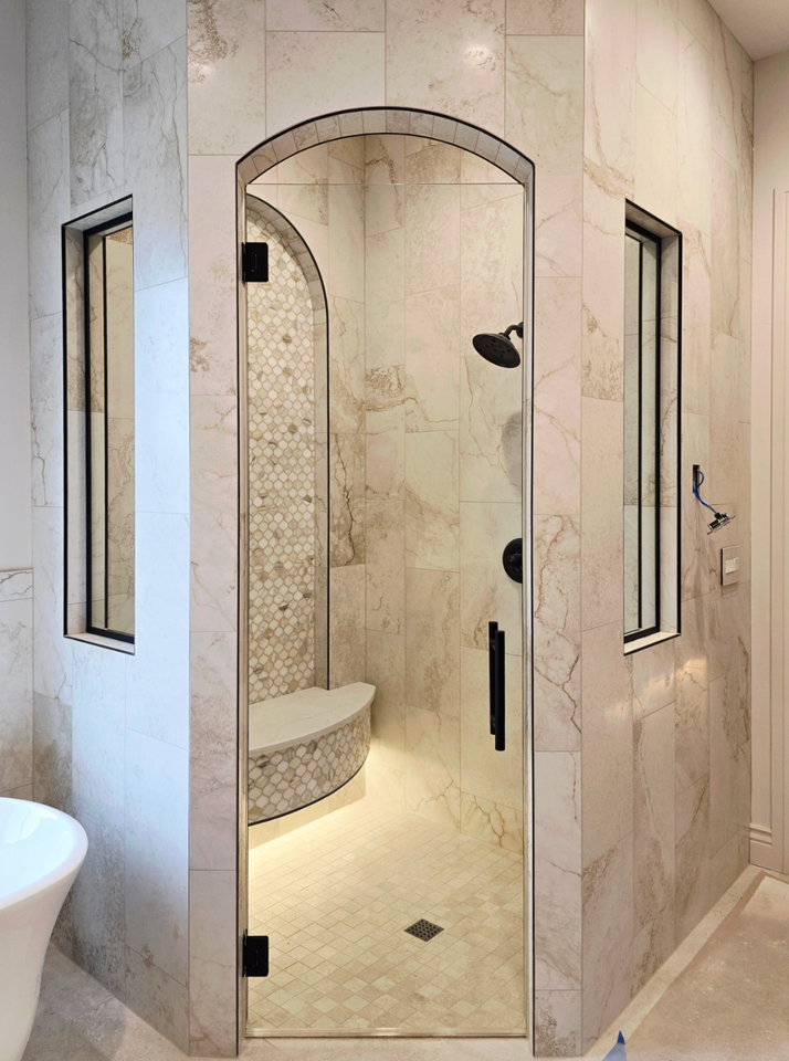 Luxurious shower with arched glass door, beige marble walls, and built-in bench.