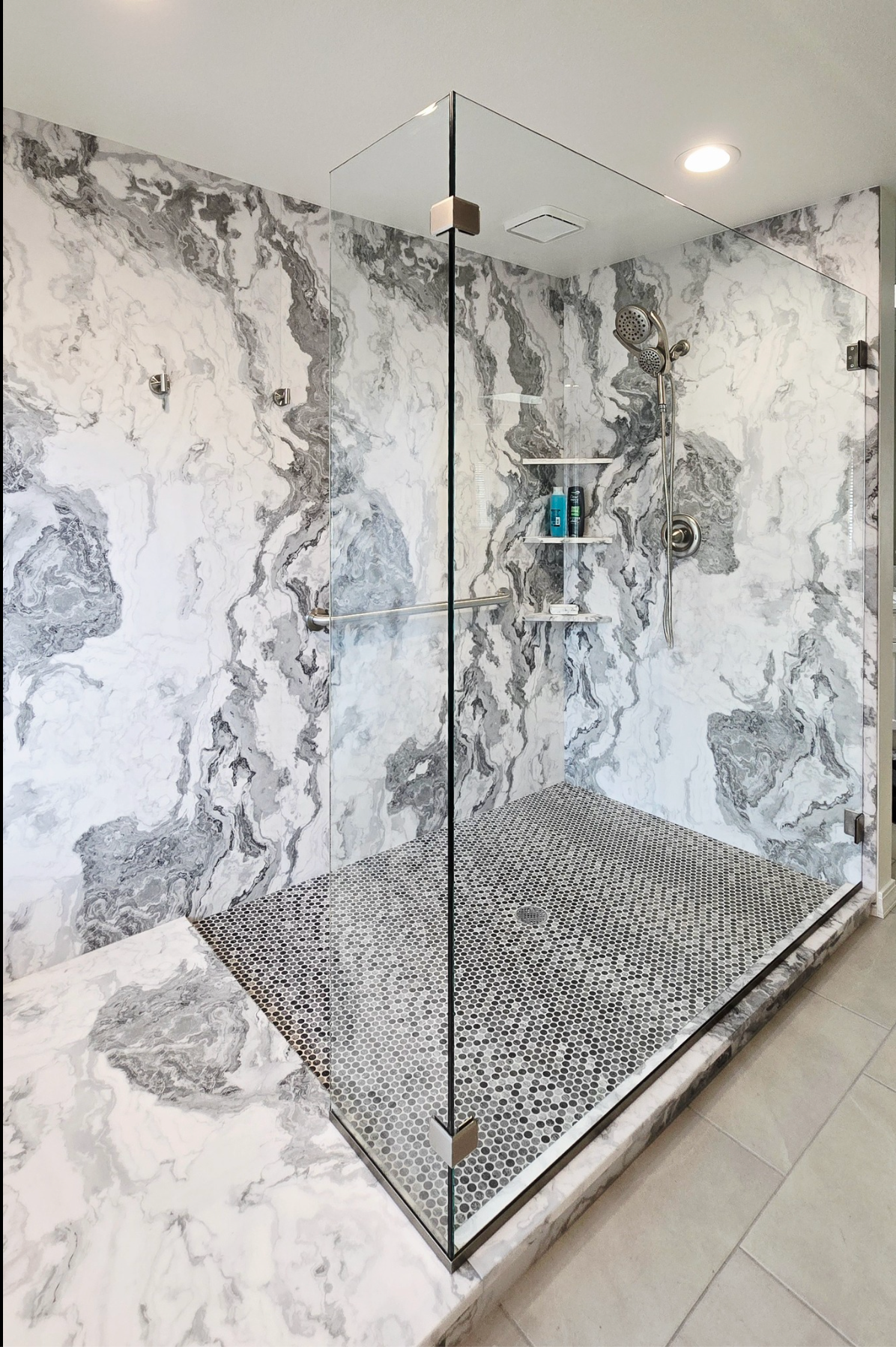 Modern shower with marble-like walls and textured floor. Glass enclosure with shower supplies on a shelf.