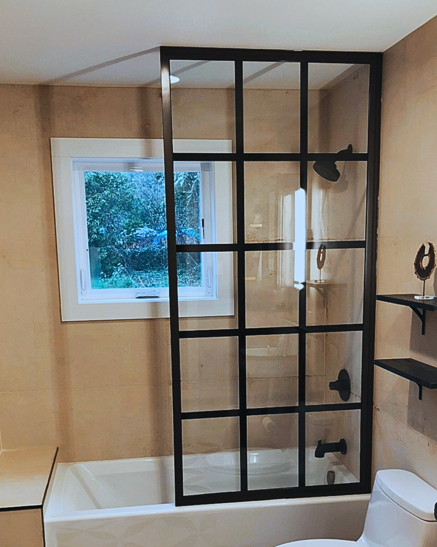 Bathroom with a black-framed glass shower screen over a white tub. Tan walls, a window, and a toilet are visible.