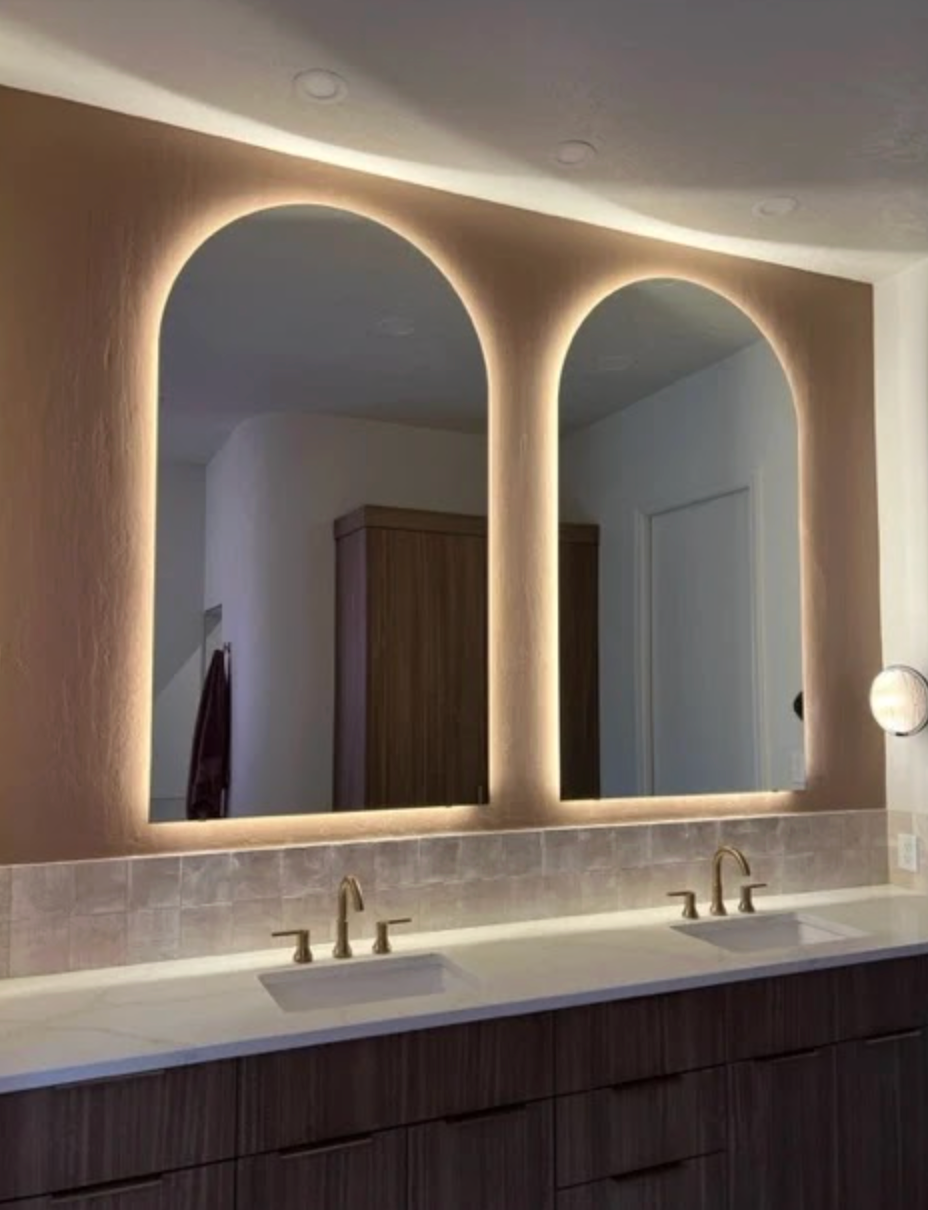 Two arched mirrors with backlighting above bathroom sinks and a vanity.