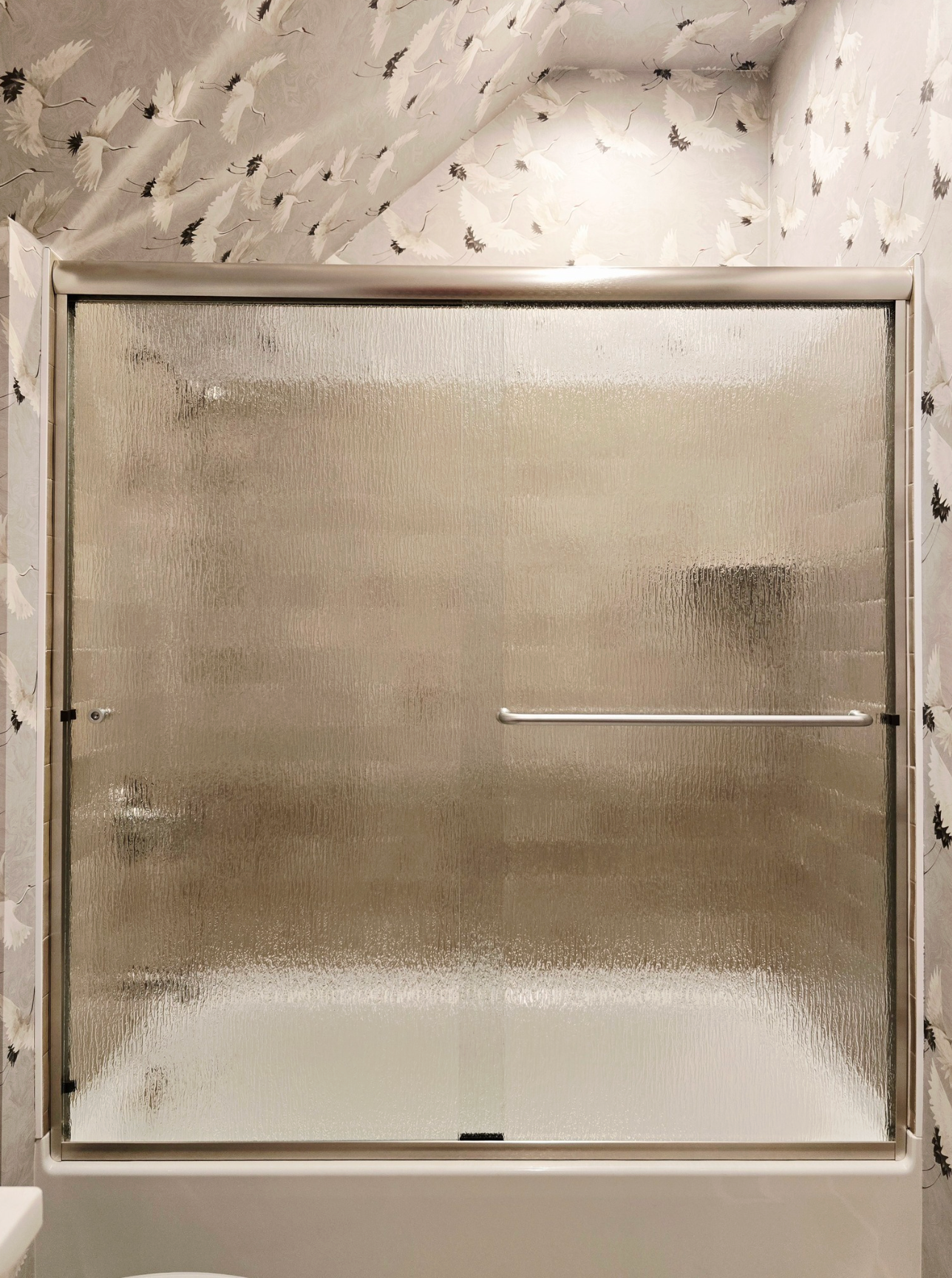 Shower with frosted glass door, silver frame, and a white bathtub. The backdrop is patterned wallpaper.