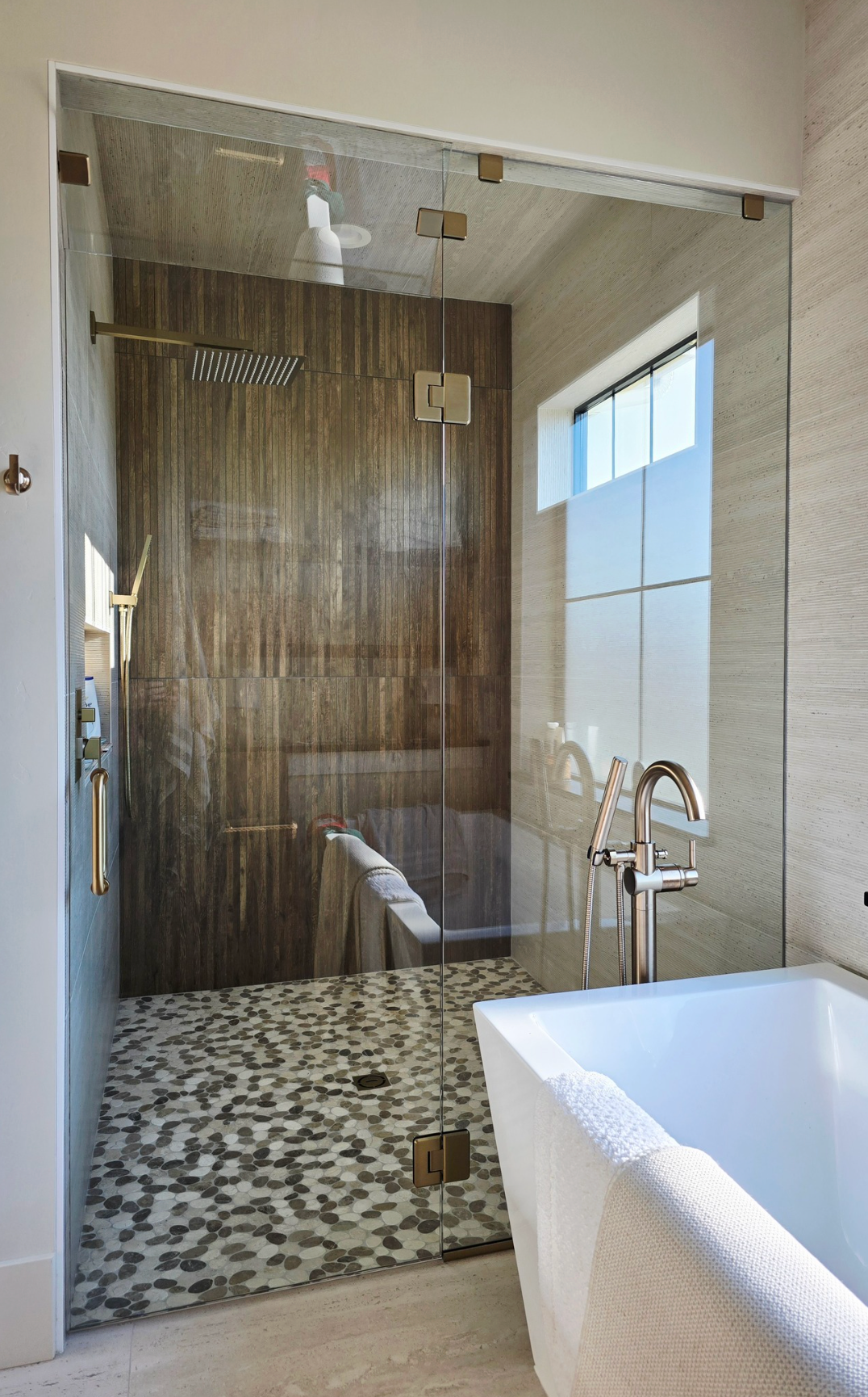 Shower with glass door, pebble floor, wood-look tiled wall, and soaking tub in a modern bathroom.
