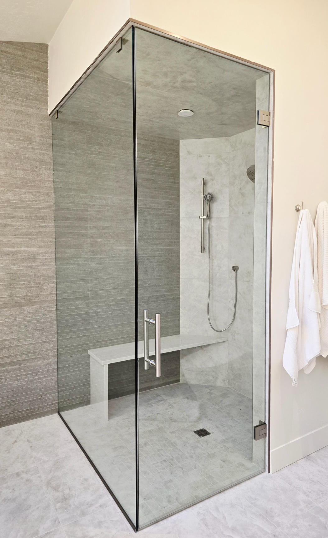 Glass-enclosed shower with a built-in bench. Gray tile interior, chrome fixtures, and glass door. White walls outside the shower.
