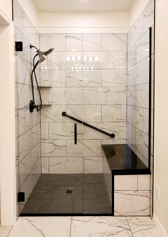 Modern shower with glass door, marble-like tiled walls, black hardware, grab bar, built-in bench.