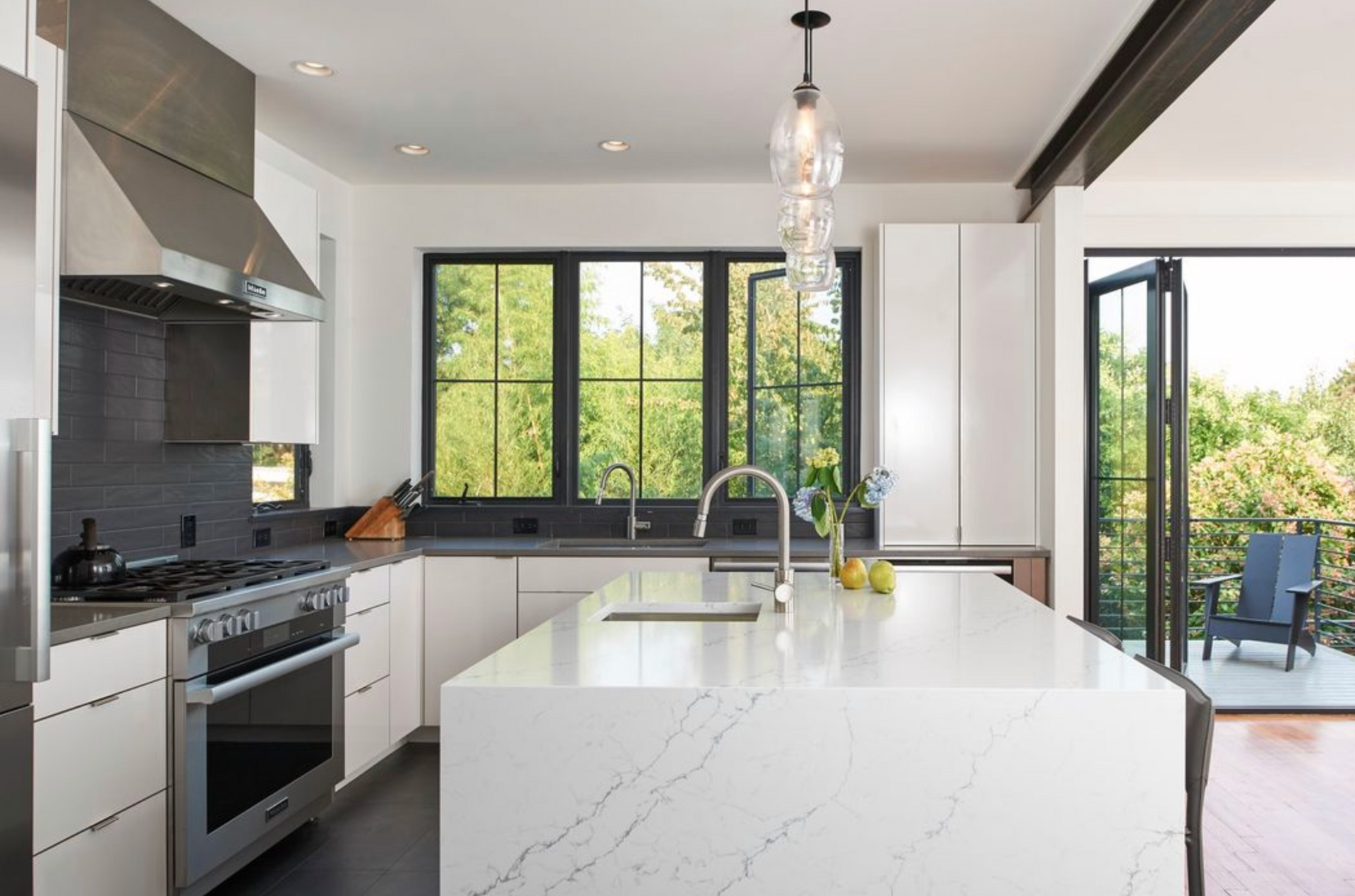 Modern kitchen with white marble island, stainless steel appliances, dark counters, and large windows looking outside.