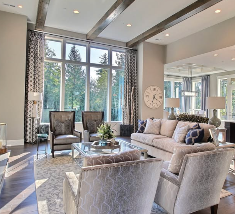A bright, modern living room with gray furniture, large floor-to-ceiling windows, wooden ceiling beams, and a wall clock.