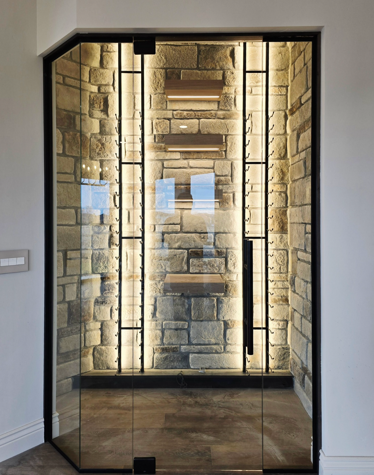 Wine cellar with glass door, stone walls, black metal accents, and interior lighting.