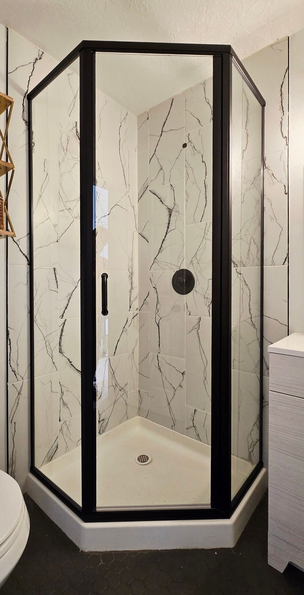 Black-framed, corner shower with patterned white walls and a white base. Black fixture.