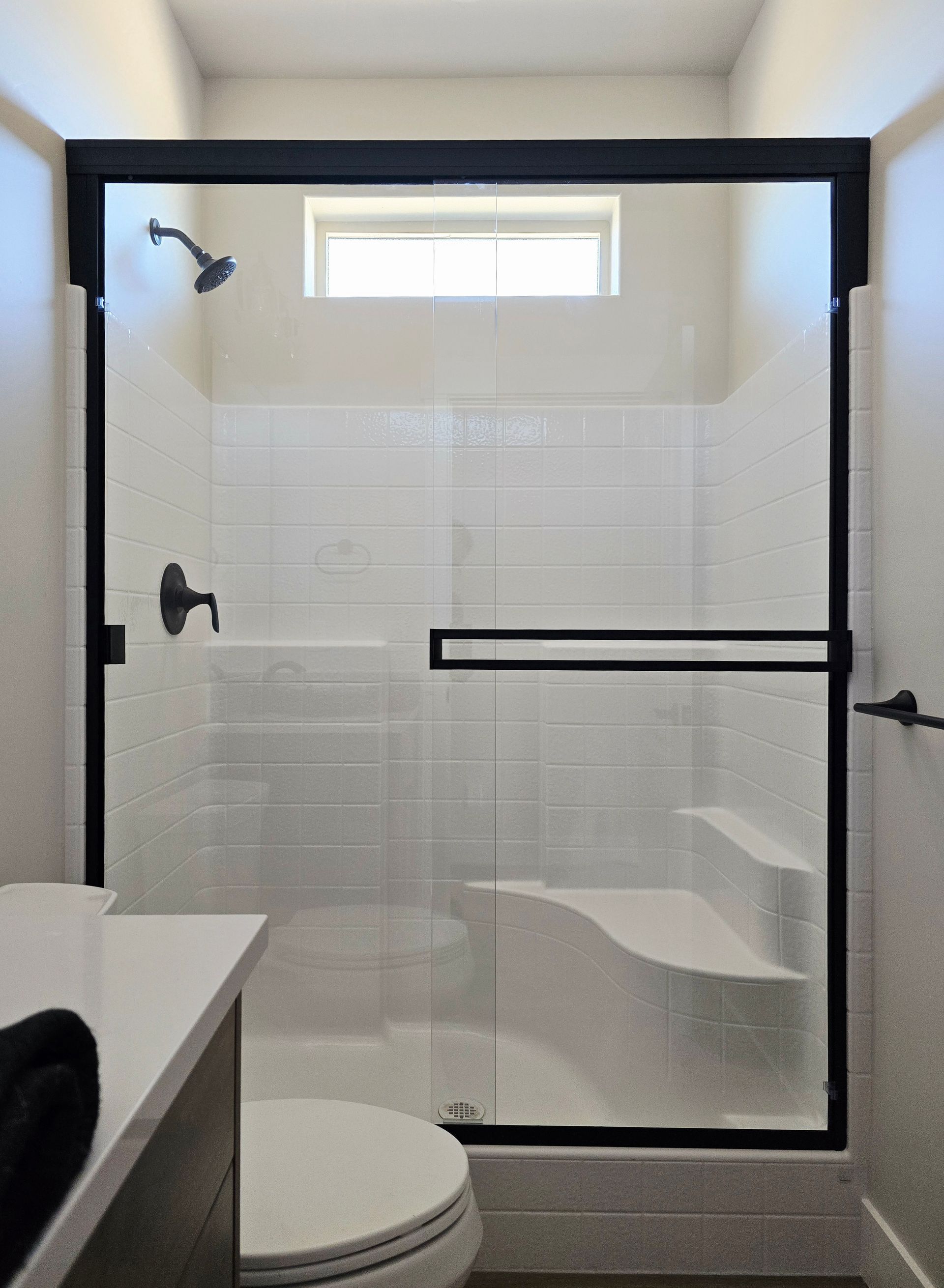 A glass shower stall with a black frame and a built-in white corner bench, viewed from a bathroom with a white vanity.