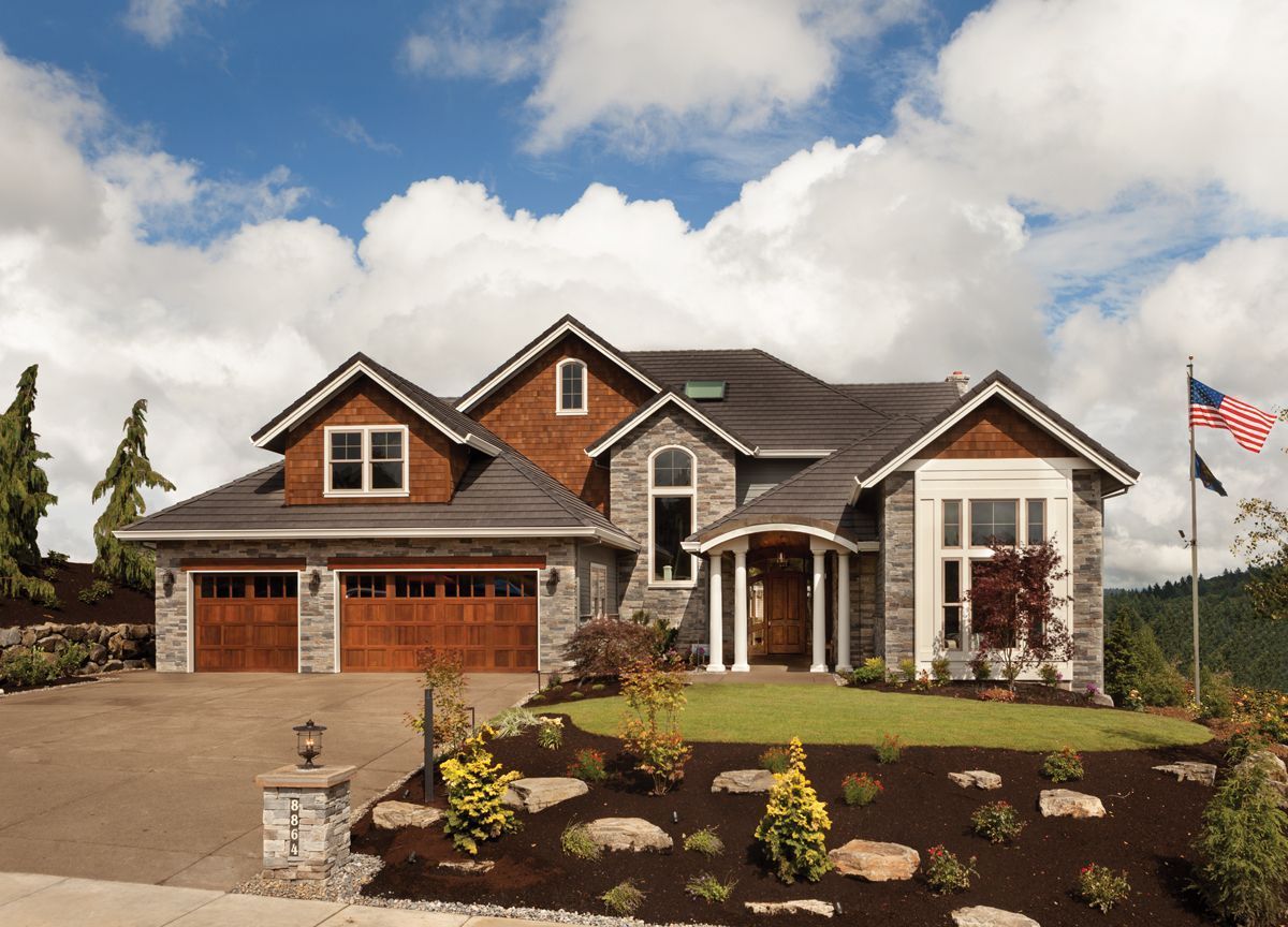 Stone and wood house with garage doors, under a cloudy sky. An American flag flies.