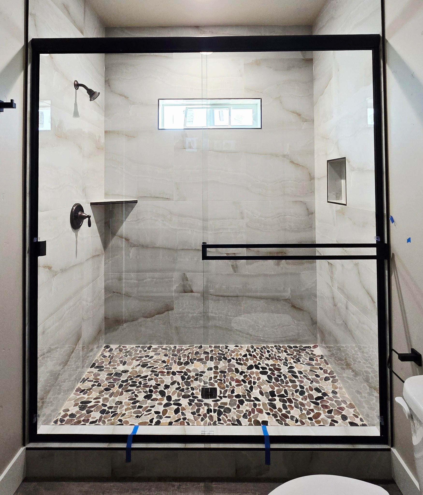 Modern shower with black-framed glass enclosure, marble-look tile, and pebble floor.