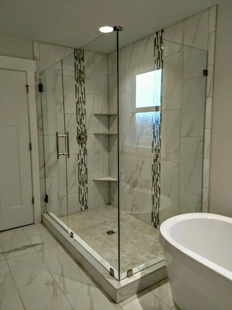 Glass shower enclosure with white and mosaic tiles. A white tub is in the foreground.