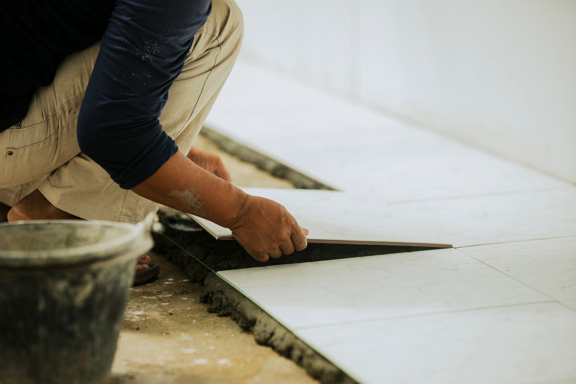 Person laying white tiles on a floor, with a bucket in the foreground.
