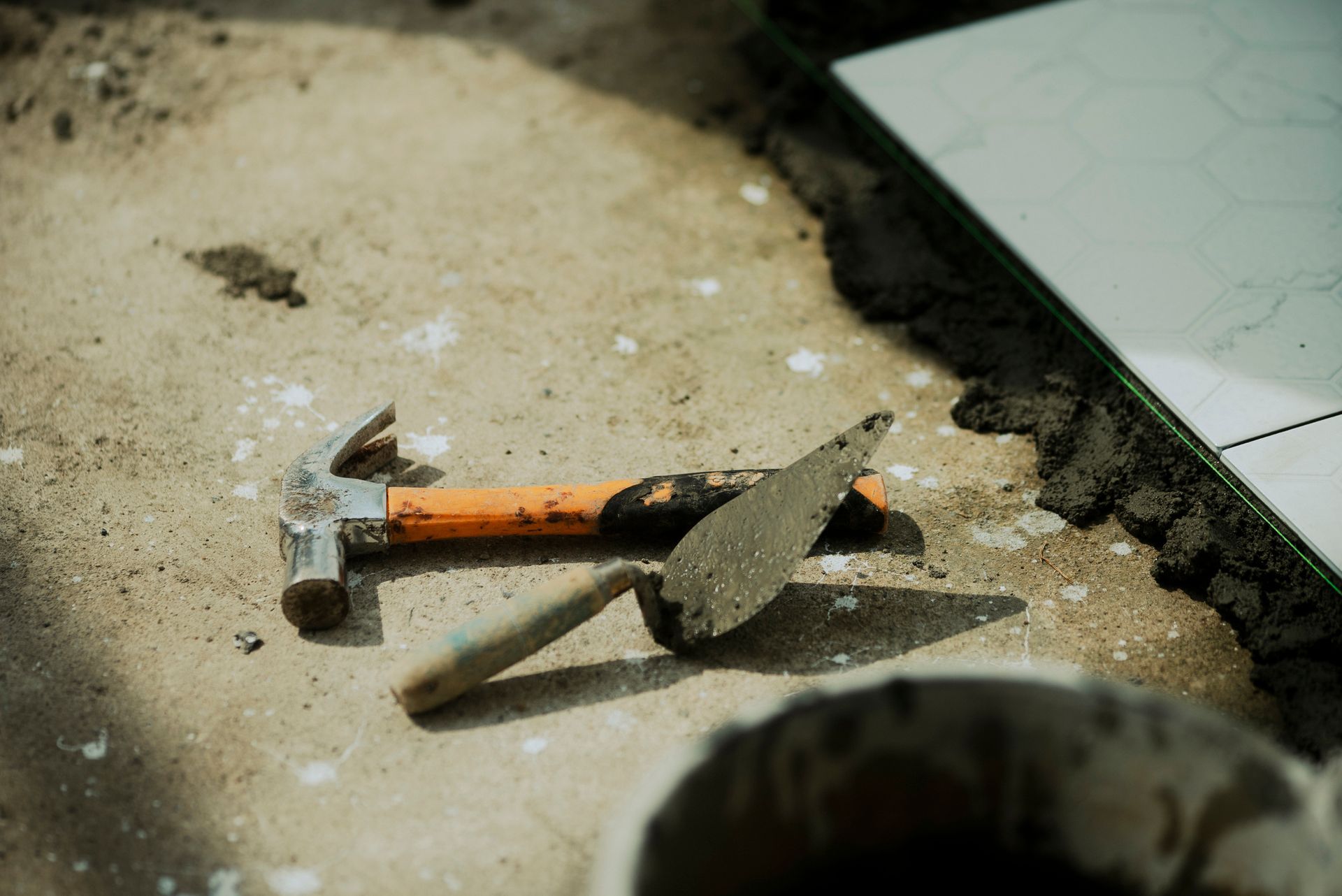 Hammer and trowel on a concrete surface next to a tiled area. Tools are orange, silver and wood.