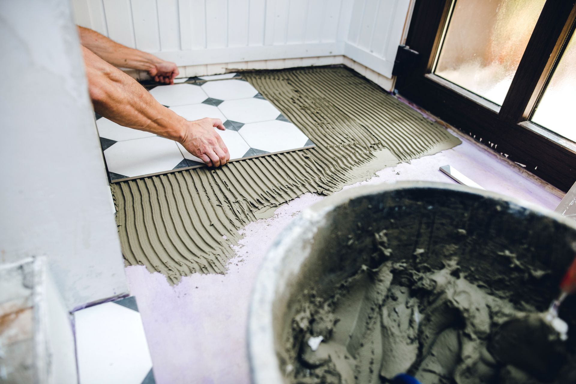 Person laying patterned floor tiles, spreading adhesive with a notched trowel. Bucket of mortar in foreground.