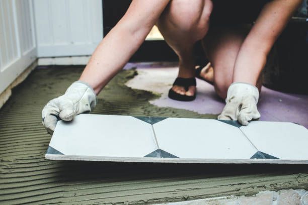 Person in work gloves installing white tiles on a floor, kneeling in a room.