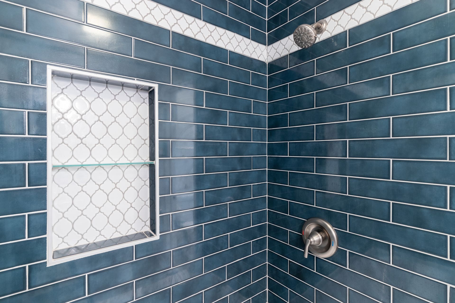 Blue tiled shower with a niche and showerhead. White decorative tile band.