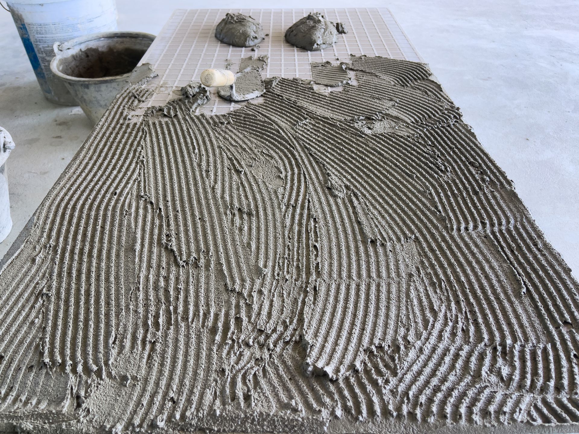 A rectangular tile surface covered in grooved mortar, with globs of mortar and a bucket nearby.