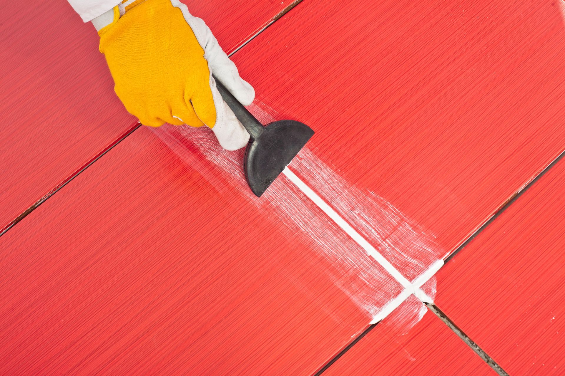 Person wearing yellow gloves applying white grout to red tile with a black trowel.