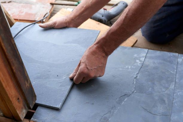 Hands laying grey stone tiles on a wooden surface outdoors, near a wooden structure.