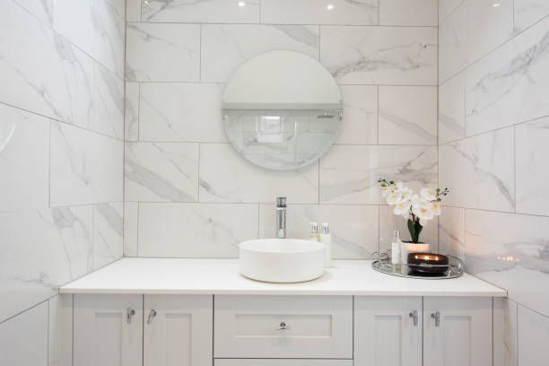 White marble-tiled bathroom with a circular mirror, white countertop, and vessel sink.