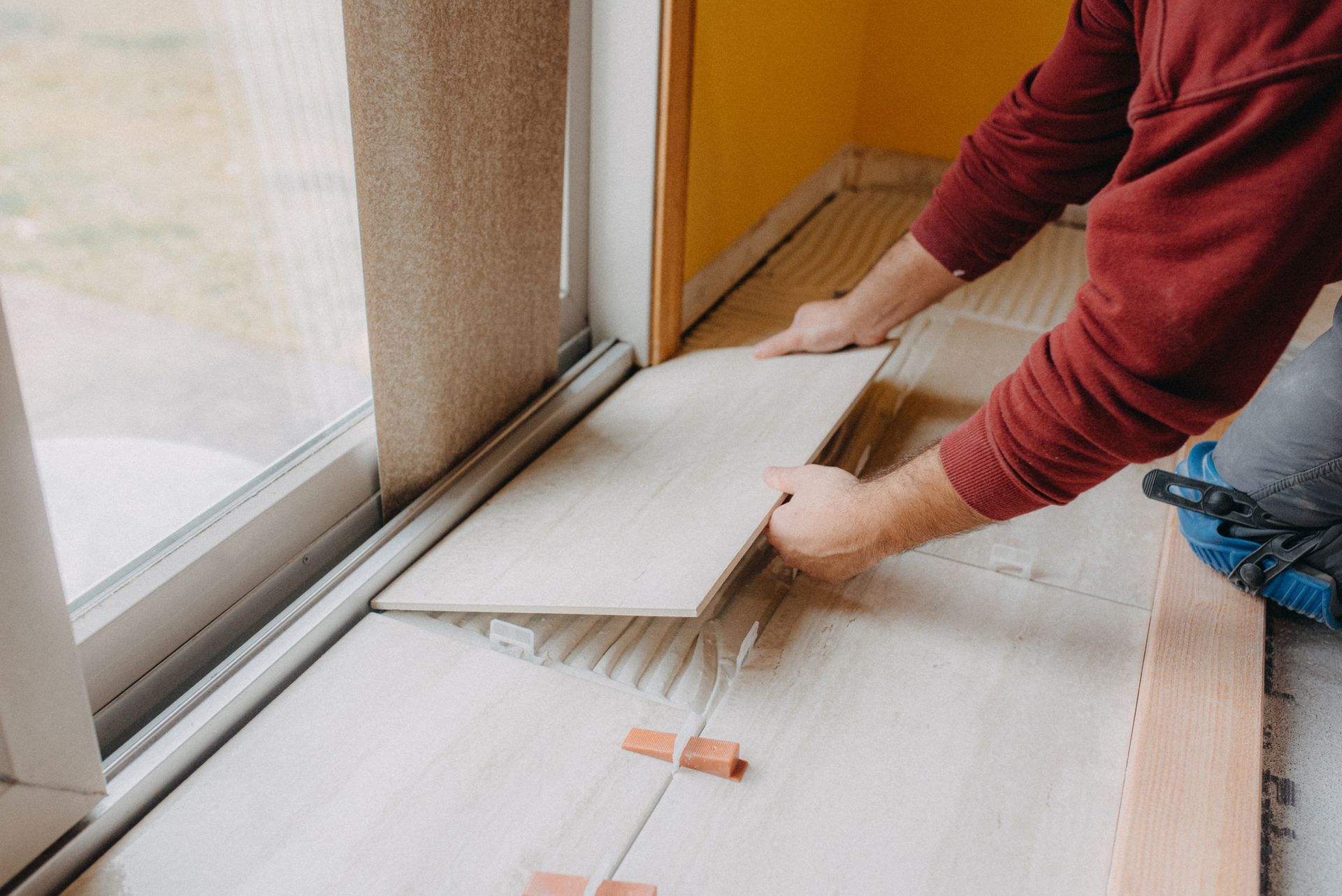 Person installing floor tile by a window. Hands placing tile on adhesive, setting in room.