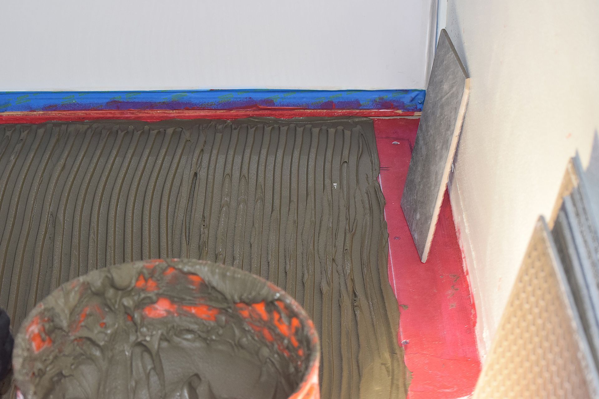 Close-up of floor with troweled mortar, bucket of mortar, and tiles being installed on a wall.