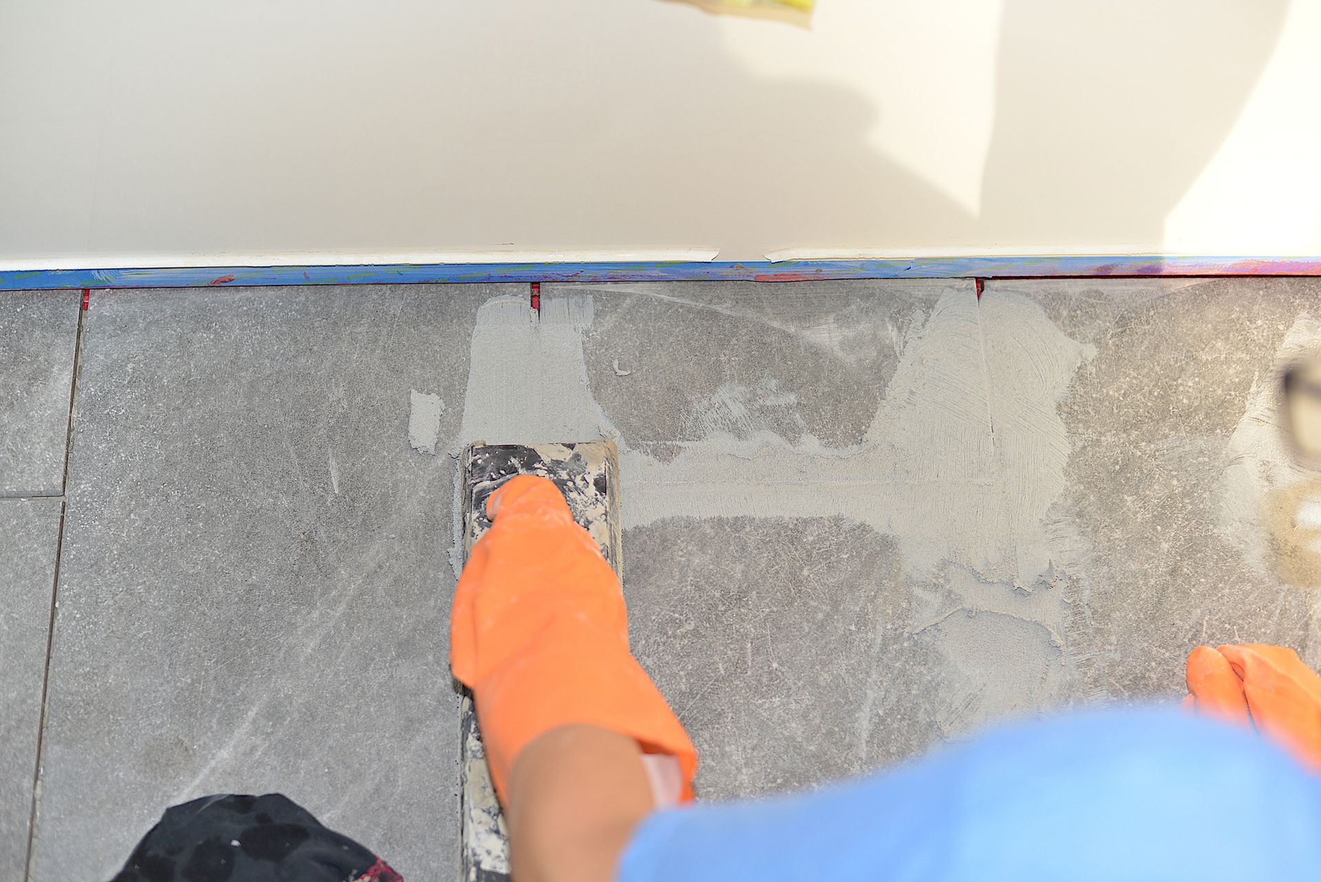 Person wearing orange gloves using a trowel to apply grout to stone tiles on a floor.