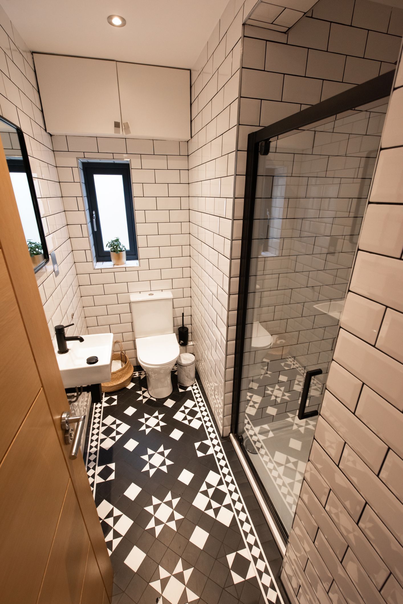 Small bathroom with black and white patterned floor, white brick-like tiles, toilet, and glass shower door.