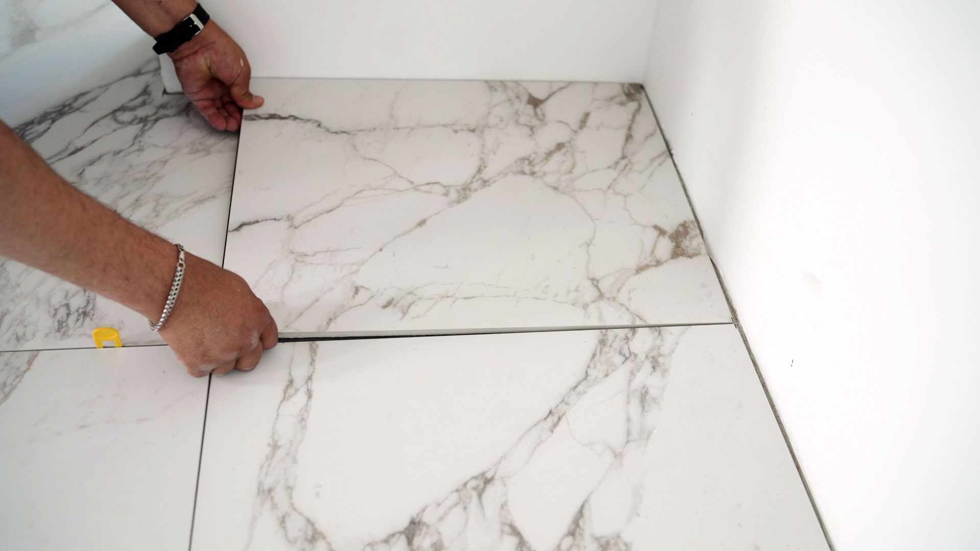 Hands placing marble-patterned tile on a floor during installation; adjacent to a wall.