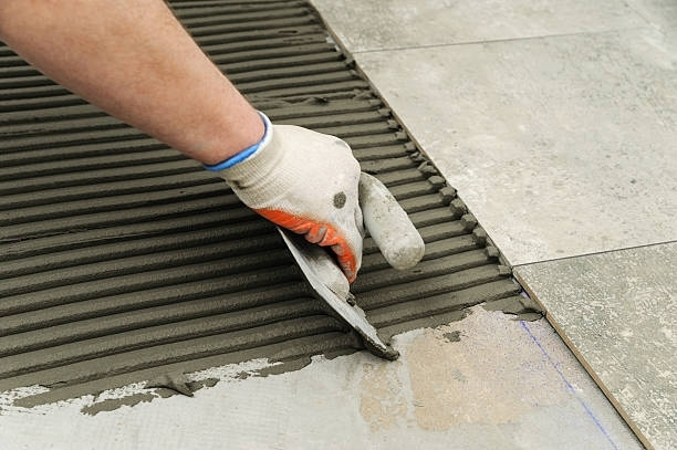 Person with gloved hand using a notched trowel to spread mortar for laying tiles.