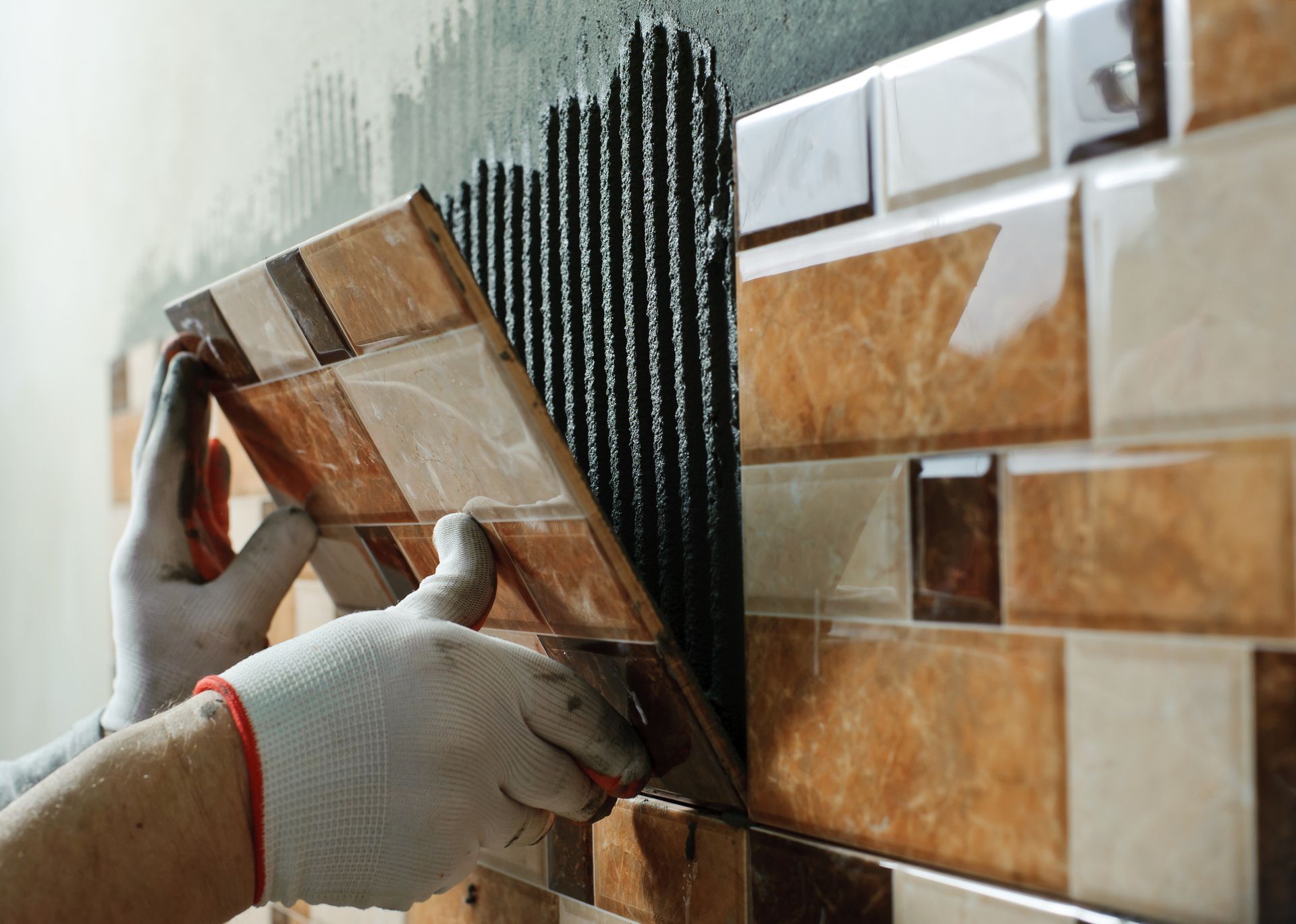 Person wearing gloves installing mosaic tile on a wall with dark adhesive.
