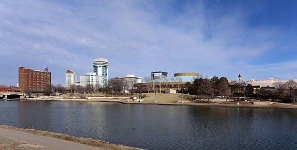 Skyline of Wichita, Kansas, reflected in the Arkansas River. Buildings, trees, and blue sky with clouds.