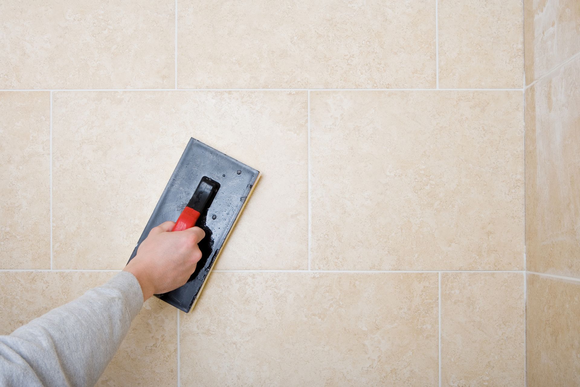 Hand with trowel applying grout to beige tiled wall.
