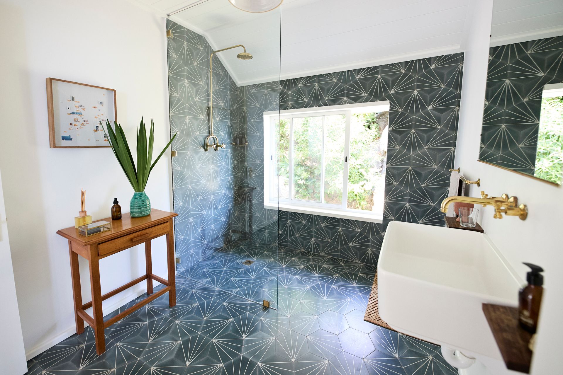 Bathroom with patterned blue-green tiles, glass shower, wooden vanity table, white sink, and window with greenery view.