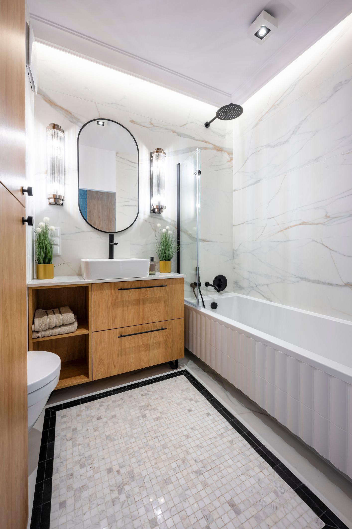 Modern bathroom with white marble walls, wood vanity, oval mirror, and patterned floor.