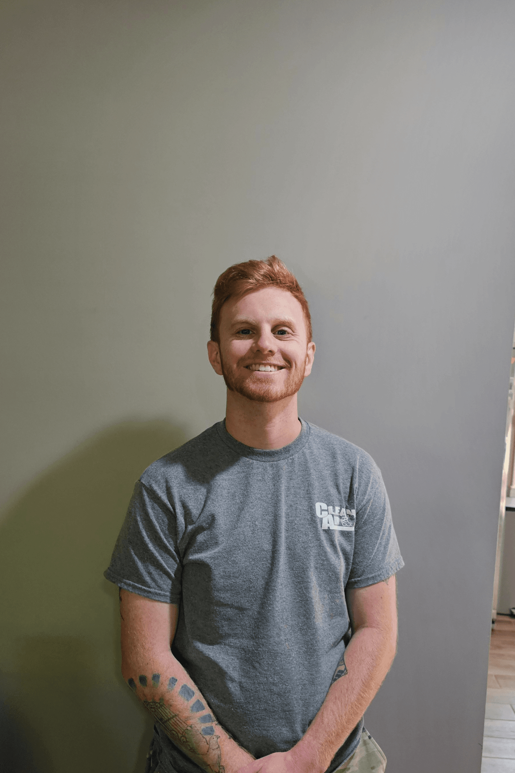 A man with red hair and a beard is standing in front of a wall with his hands behind his back.