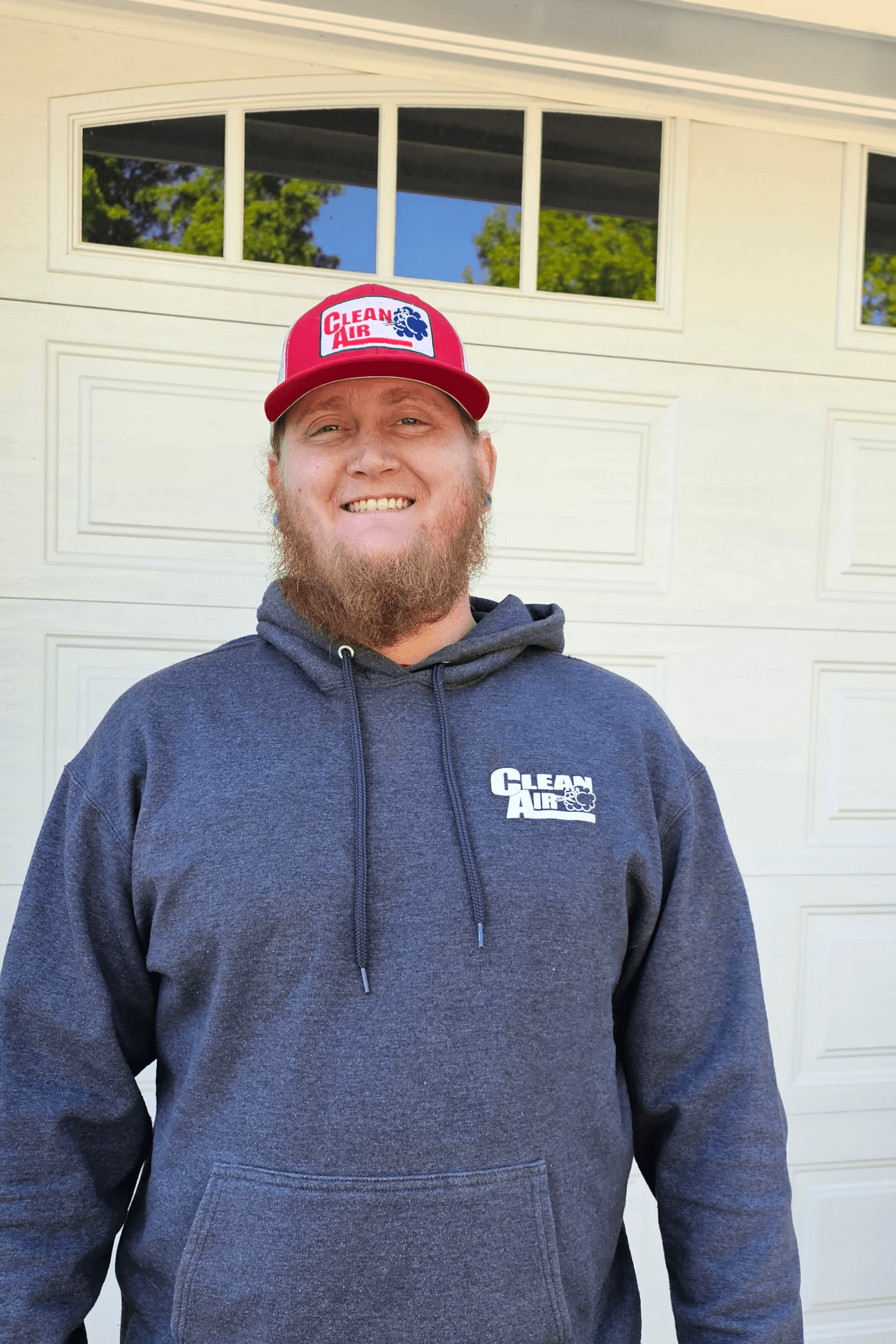 A man with a beard wearing a red hat and a hoodie is standing in front of a garage door.