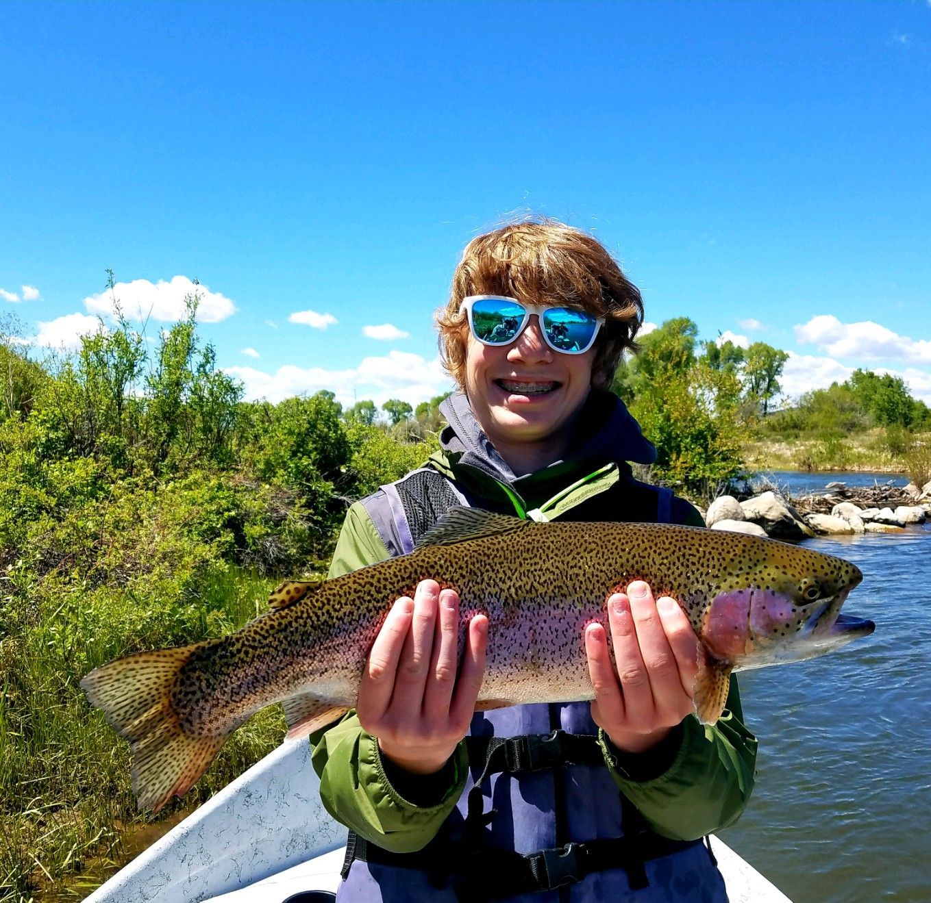 Person on a boat holding a large fish, smiling on a sunny riverbank