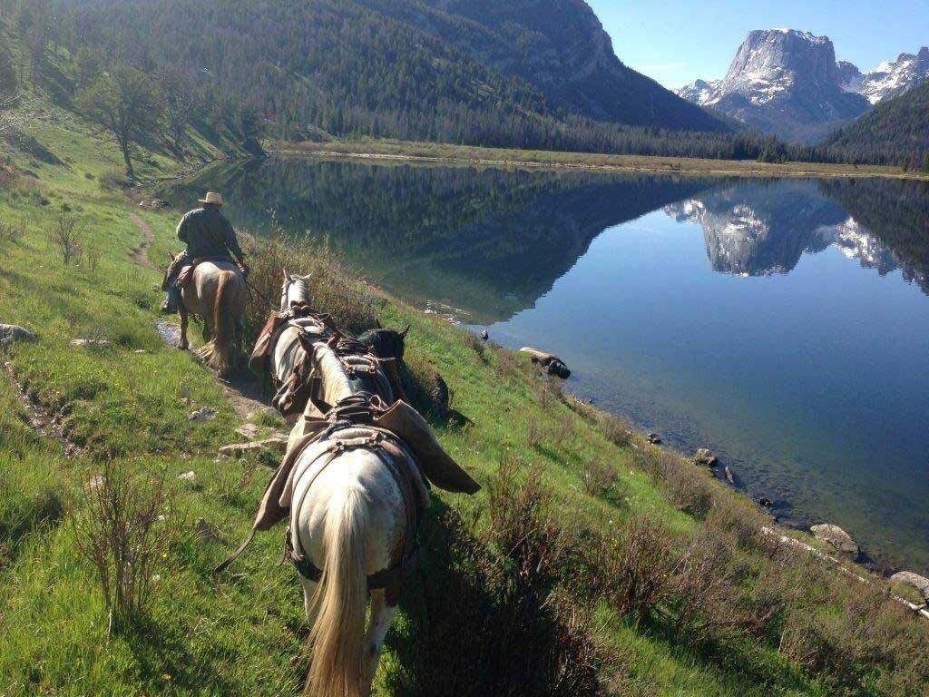 Riders on horseback follow a lakeside trail beside a calm mountain lake reflecting peaks and sky.