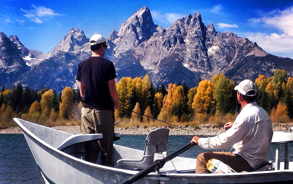 Two people fishing from a boat on a mountain lake with autumn trees and rocky peaks in the background