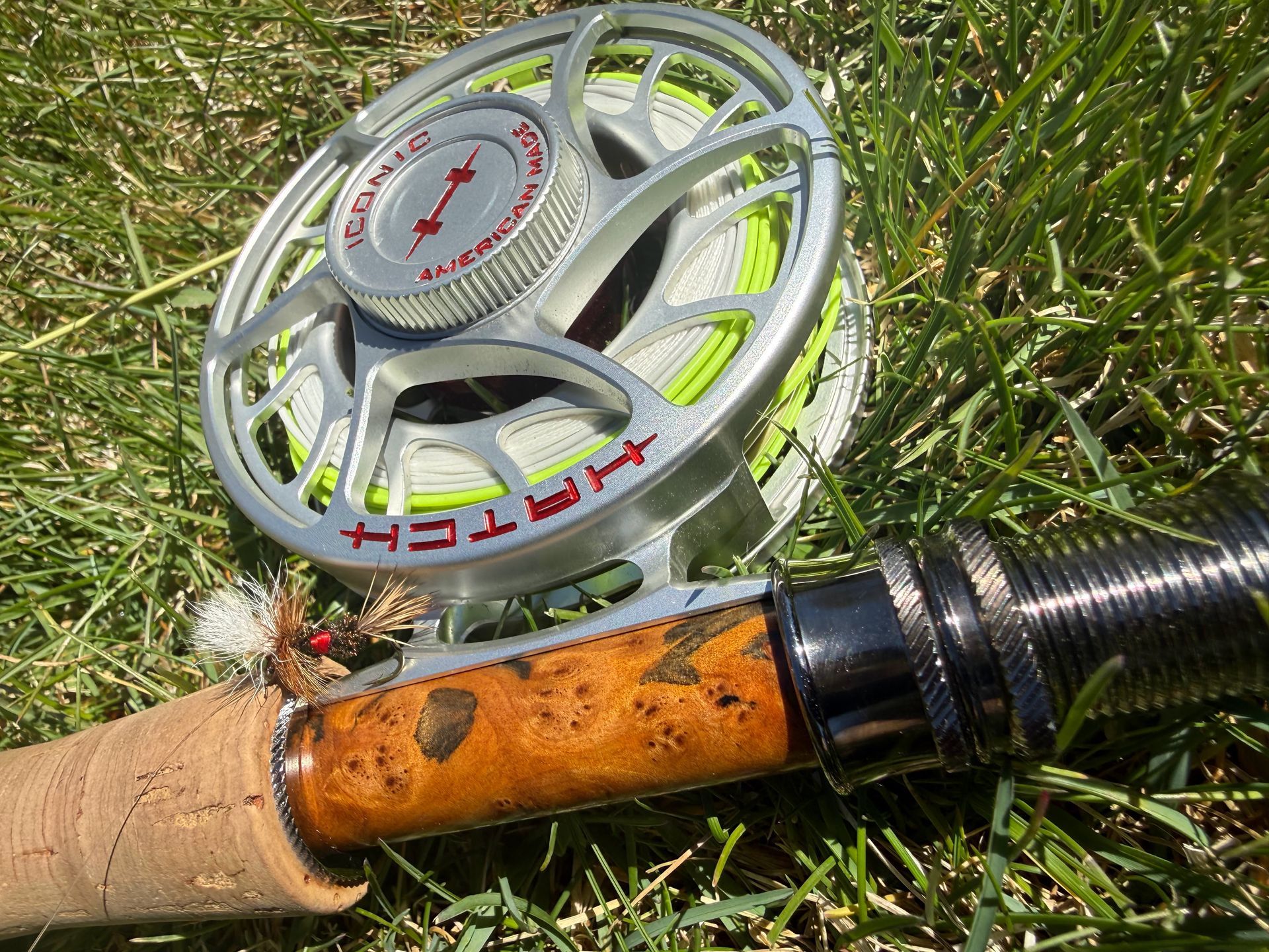 Fly reel on a fishing rod resting in grass, with orange and silver details.